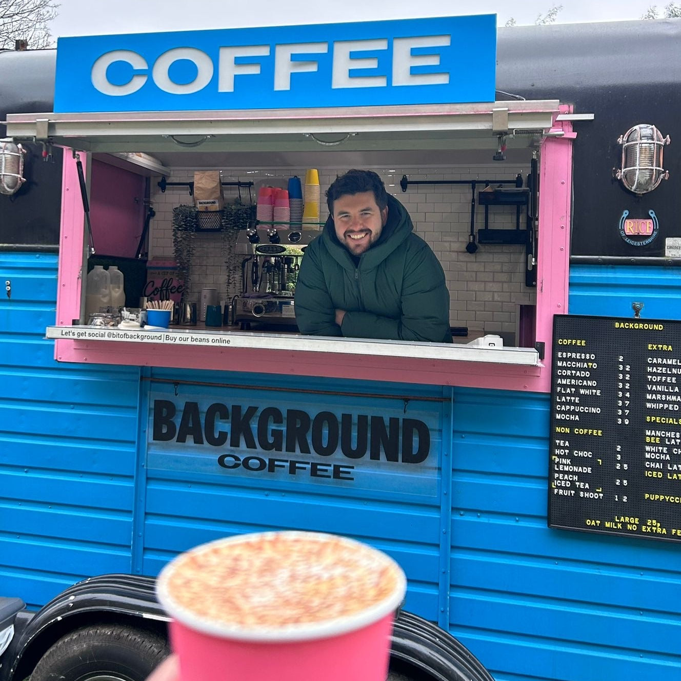 Person standing behind a coffee truck with a customer holding a pink cup.